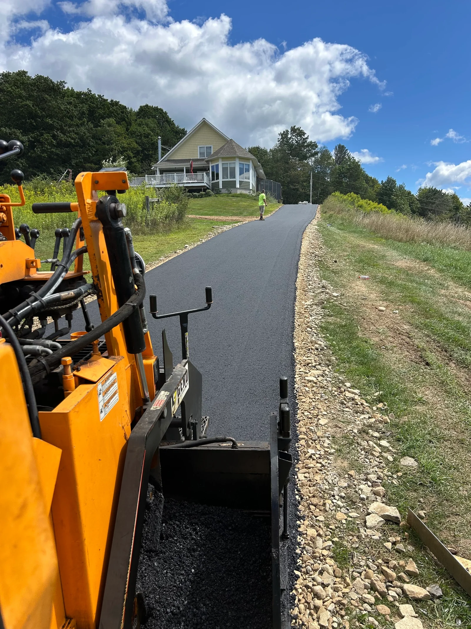 Newly paved asphalt driveway with stone base and residential home in background