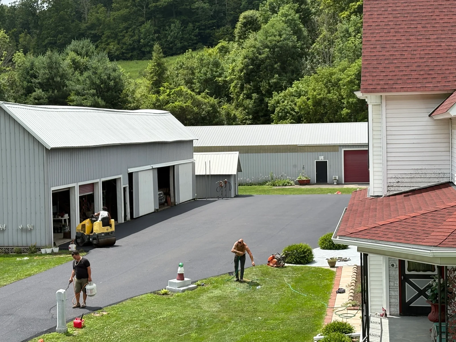 Freshly paved asphalt driveway with metal storage buildings and residential home in background